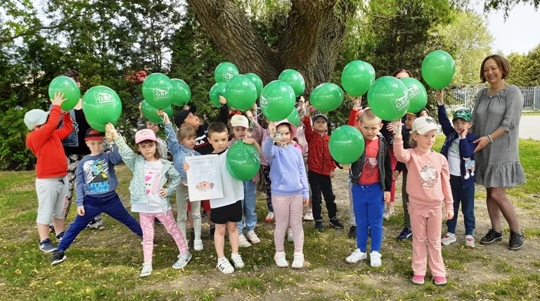 Na zdjęciu znajdują się dzieci z zielonymi balonami z logotypem Gminnej Biblioteki Publicznej w Zbuczynie wraz z pracownikami Biblioteki oraz nauczycielkami. Wszyscy stoją na zewnątrz pod wierzbą.